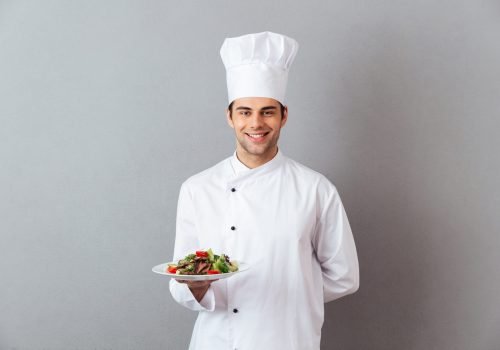 Picture of happy young cook in uniform standing isolated over grey wall background. Looking camera holding salad.