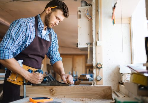 Profile view of confident young carpenter in apron planing board with jointer plane in spacious workshop